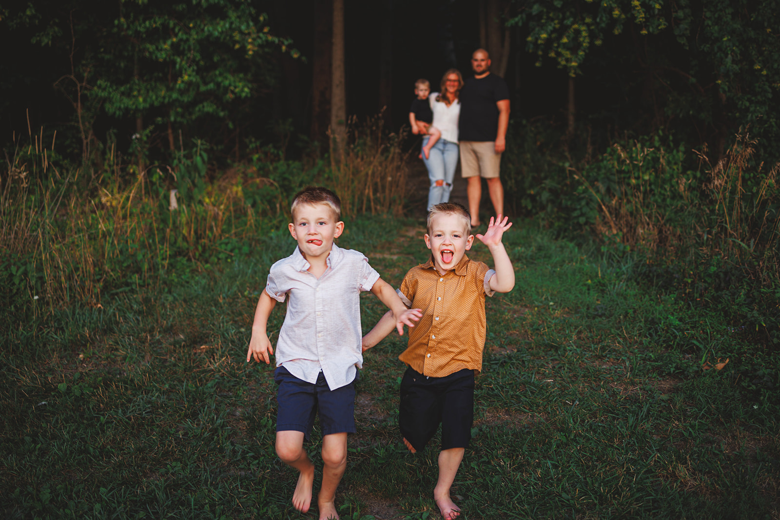 Two boys running barefoot through evening grass while their parents follow behind