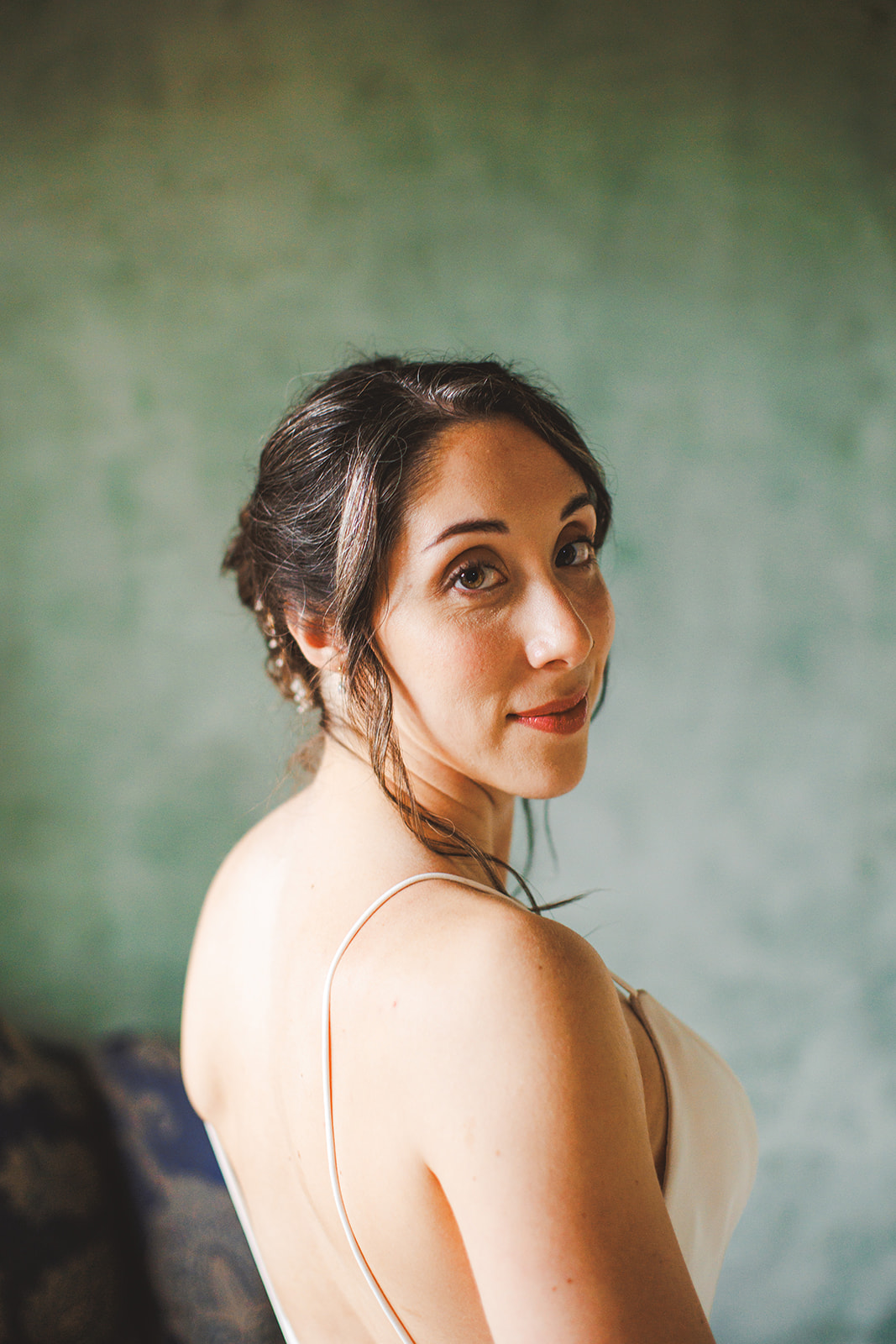 Close portrait of Amie looking back over her bare shoulder against a sea-green plaster wall, loose curls framing her face