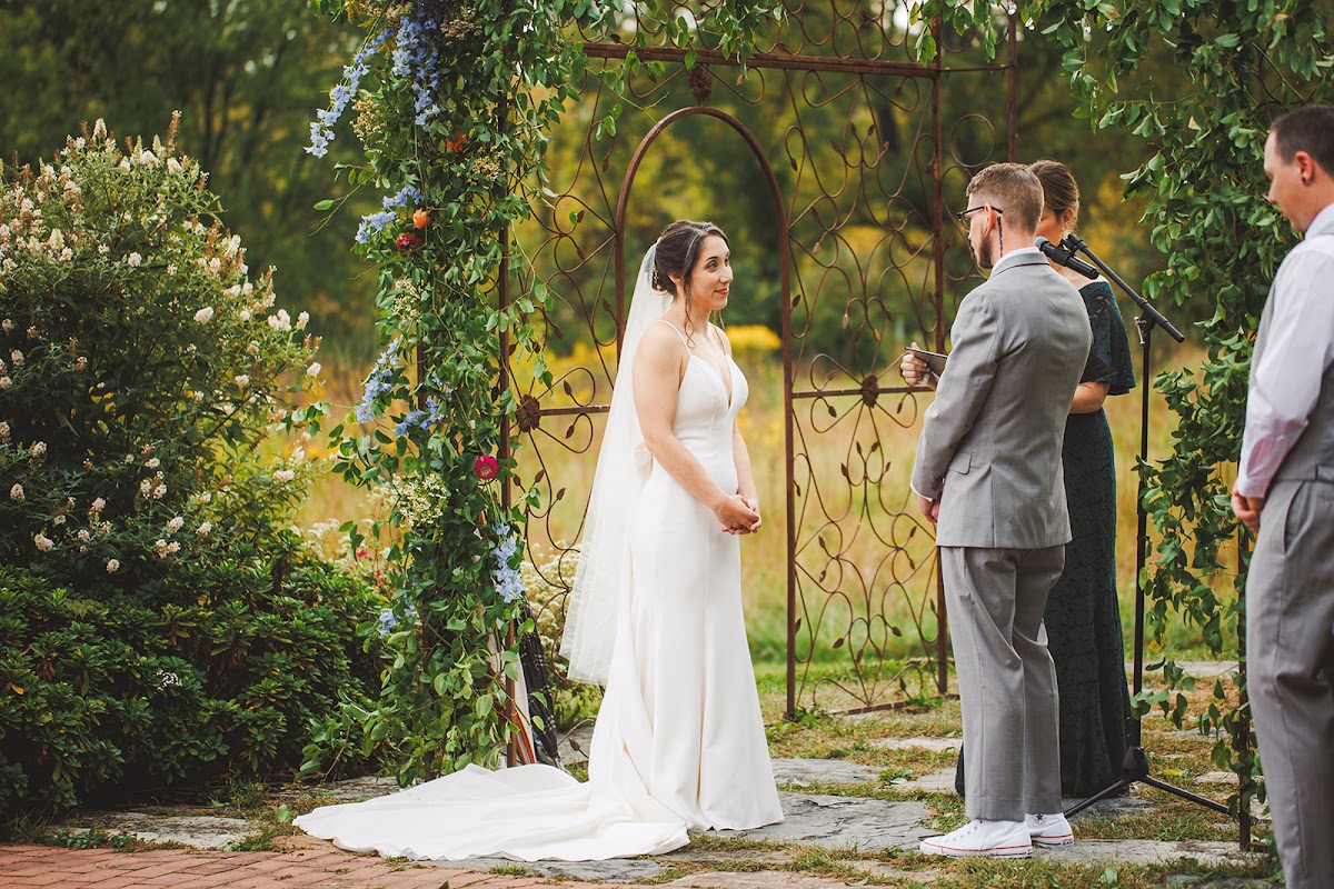 Amie standing at an iron garden arbor during her open-air ceremony, wildflower meadow and trees behind her