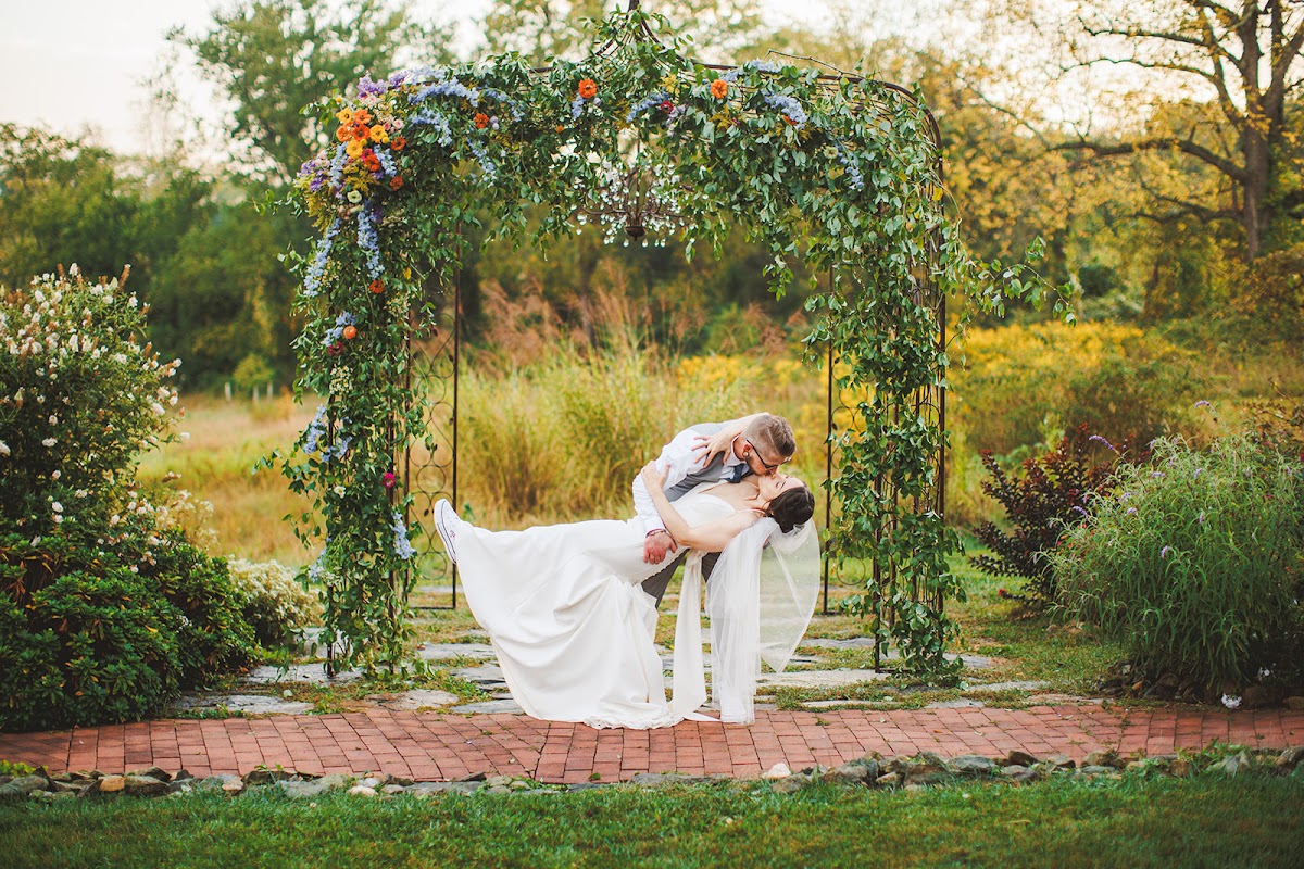 A dip kiss under a flower-laden iron arbor with the veil cascading and a golden meadow behind