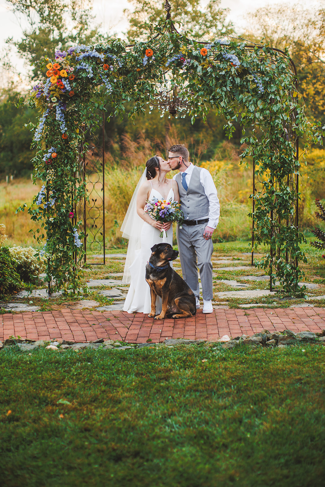 Amie and Cody kissing under the flower-draped iron arbor at golden hour while Cujo sits square in front of them on the brick path