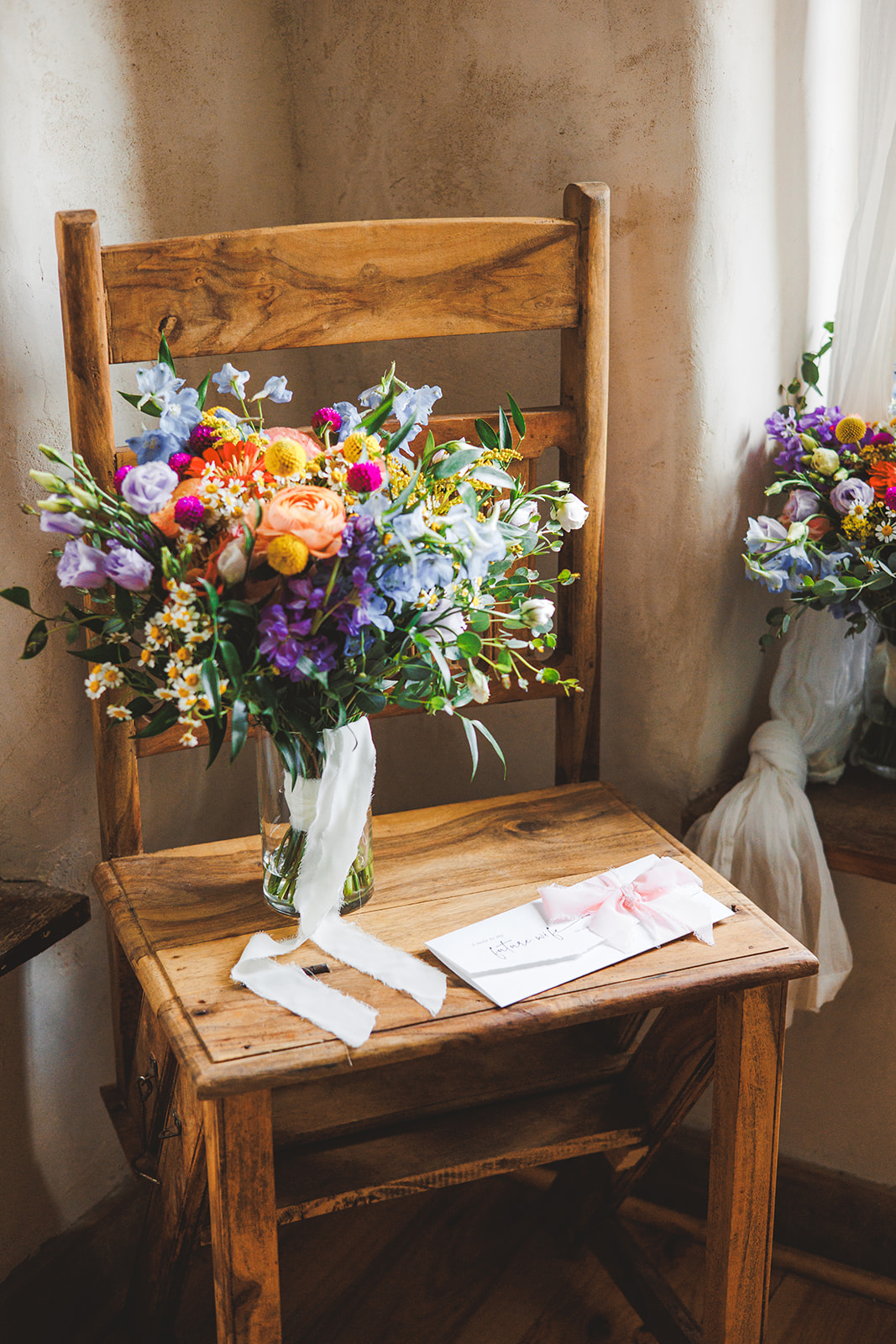 A wildflower bouquet in blues, purples, oranges, and yellows resting on a wooden chair beside a card tied with a pink bow addressed to the future wife