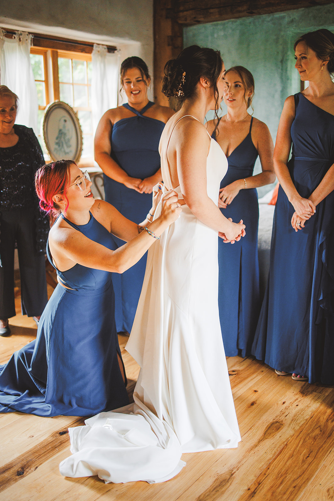 A bridesmaid in blue kneeling to fasten the back of Amie's dress while the rest of the party watches in the timber-framed farmhouse room