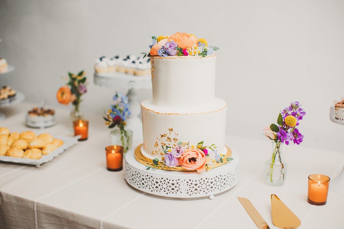 Two-tier white wedding cake with gold leaf and fresh flowers in orange, lavender, and blue on the dessert table, surrounded by bud vases and candles