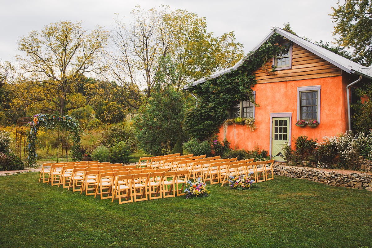 Rows of wooden folding chairs on the lawn facing the ivy-covered orange cottage at Zigbone Farm Retreat, with wildflower arrangements marking the aisle