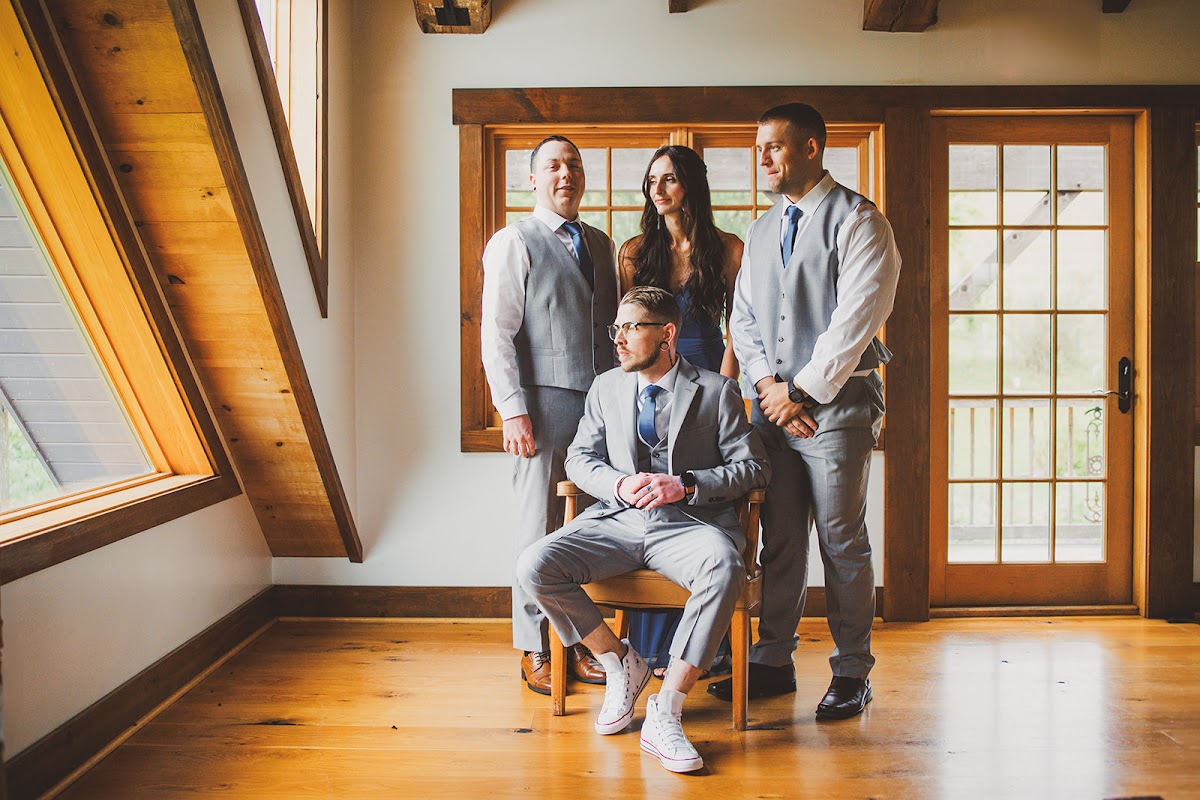 Cody seated in a chair in white high-top sneakers with his wedding party in gray vests and a navy dress standing around him in the bright loft