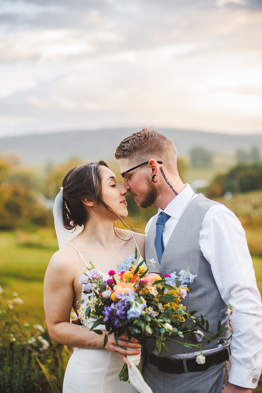 A couple resting forehead to forehead over a bright bouquet beneath a painterly sunset sky