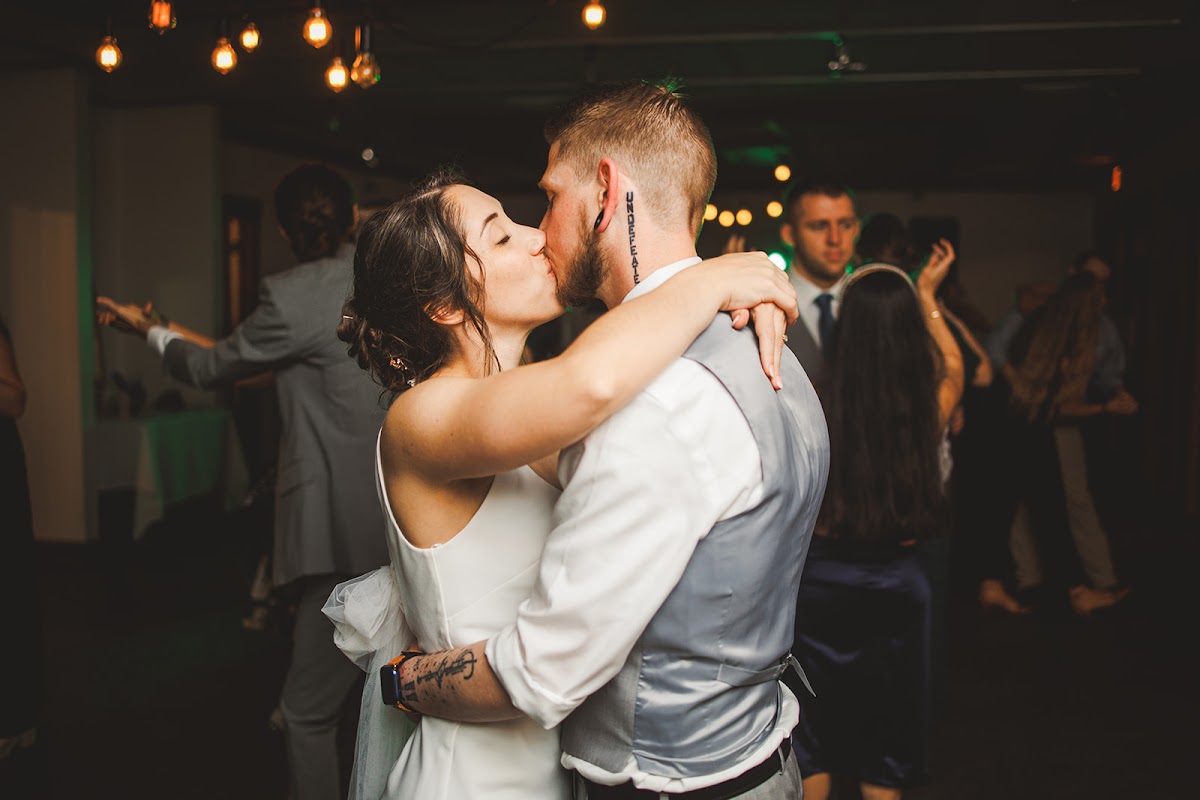Amie and Cody kissing during their first dance under cafe lights, her arms around his neck, guests soft in the background