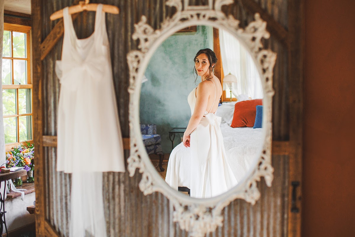 Amie reflected in an ornate white oval mirror adjusting her dress, a second white dress hanging by the window against a corrugated metal wall