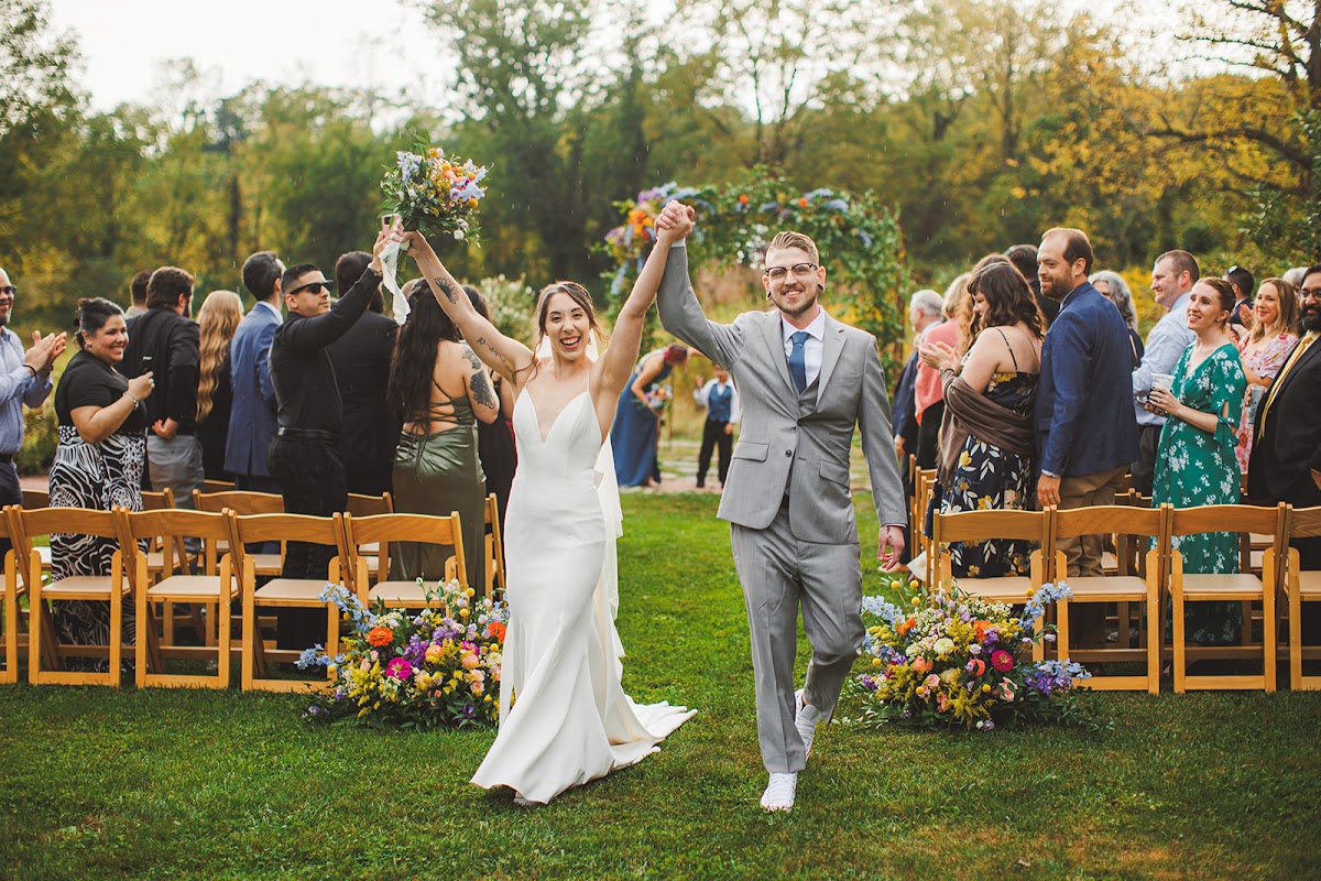Amie and Cody charging up the grass aisle with joined hands thrown in the air, the flower-covered arch and applauding guests behind them