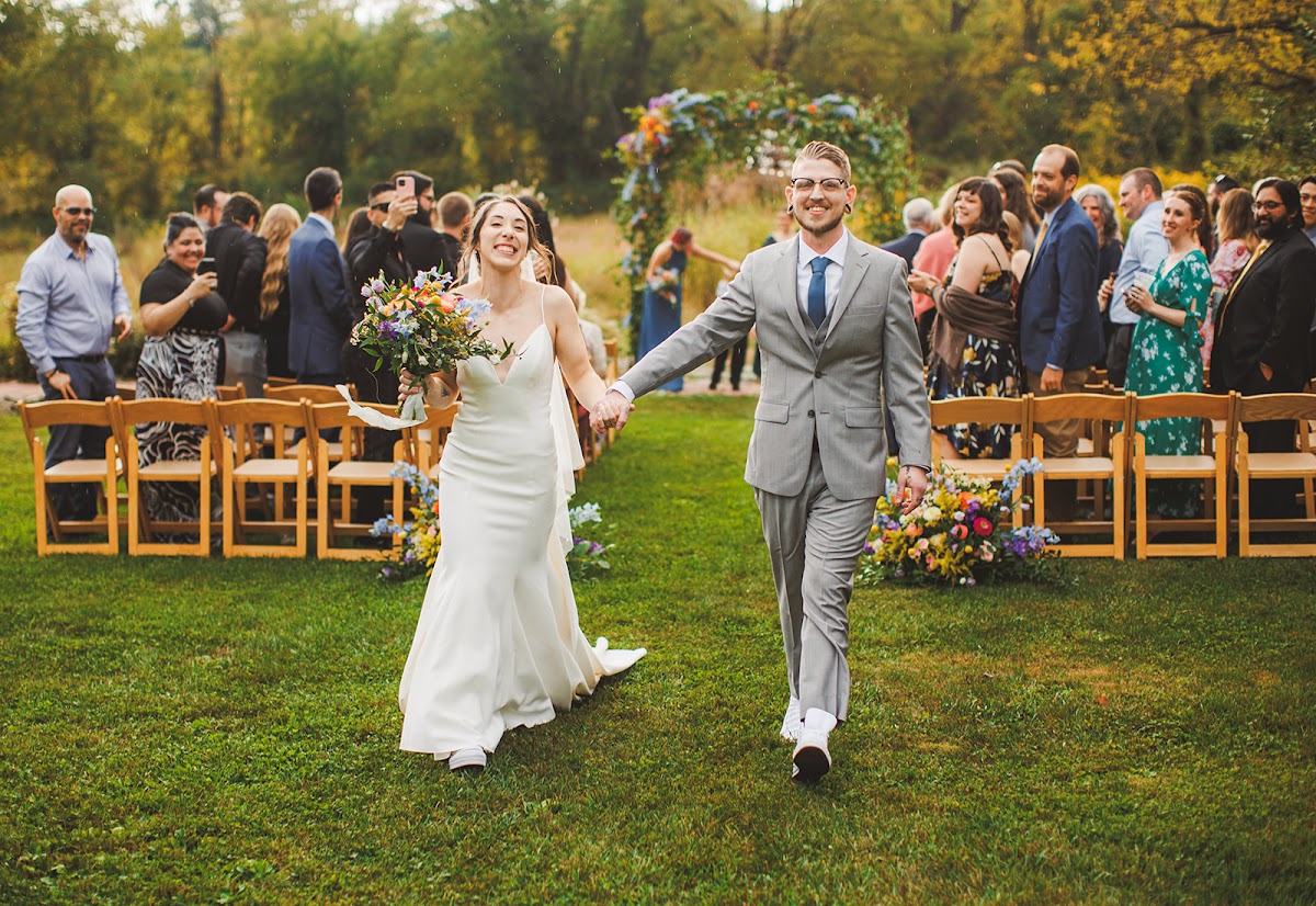 Amie and Cody walking back up the grass aisle after the ceremony, both grinning, her wildflower bouquet raised and guests standing on both sides