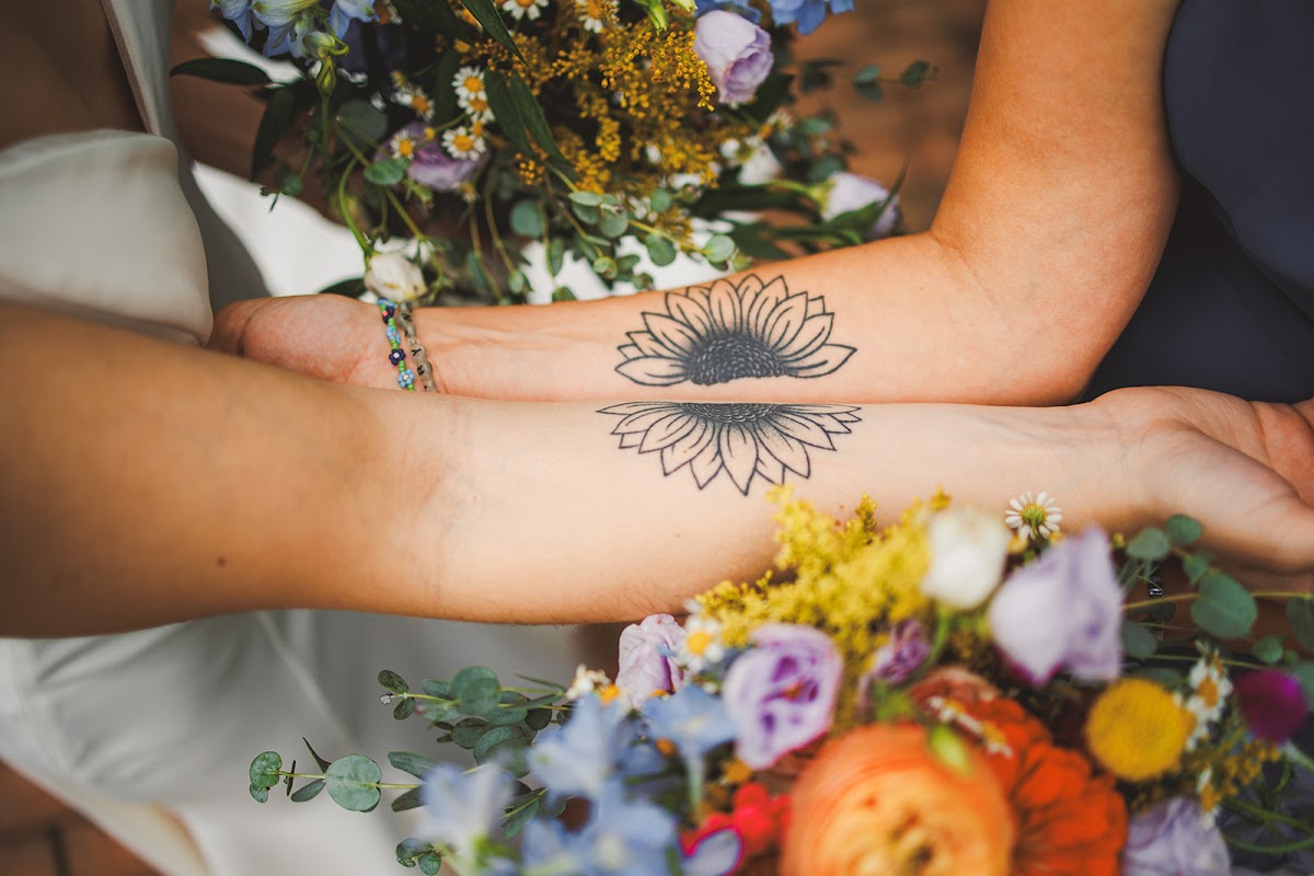 A close crop of a couple's matching sunflower forearm tattoos resting over their wildflower bouquets