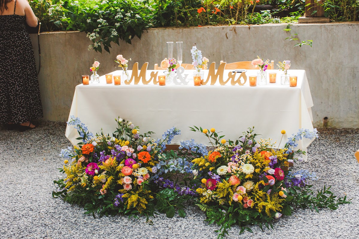 The sweetheart table at Zigbone Farm Retreat with gold Mr and Mrs letters, candles, and a dense band of colorful flowers along its base, set against a stone garden wall