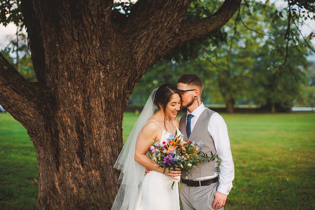 Cody kissing Amie's cheek beside a massive tree trunk, her veil catching the light and her multicolor bouquet held low against the open lawn