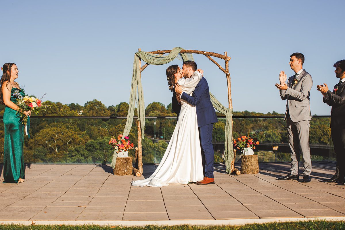 Andrea and Patrick kissing beneath the sage-draped branch arch on the Bavarian Inn terrace, the wooded Potomac valley behind them and groomsmen applauding