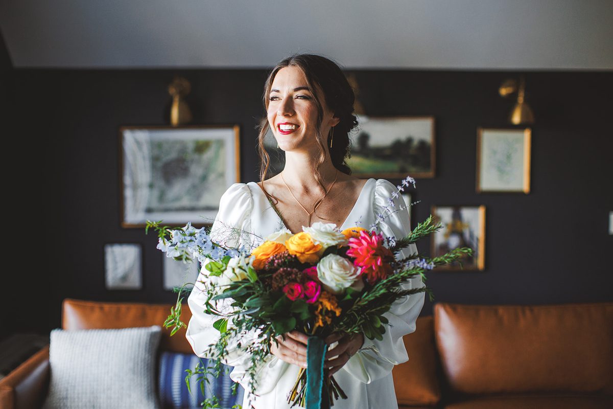 Andrea smiling with her vivid bouquet of dahlias, ranunculus, and roses in the dark-walled lounge