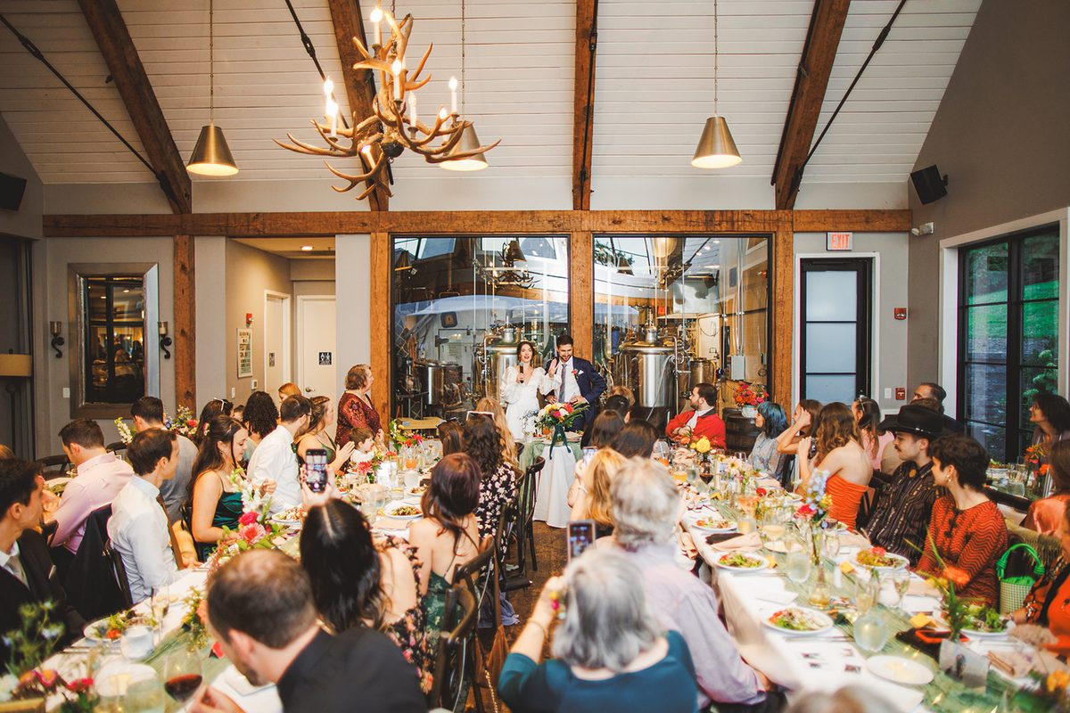 Andrea and Patrick welcoming their guests at the head of the timber-framed brewhouse room, brew tanks behind glass and an antler chandelier above