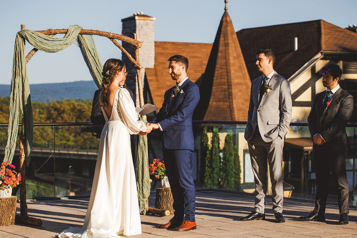 Andrea and Patrick holding hands beneath the arch during the ceremony, the Blue Ridge soft in the distance