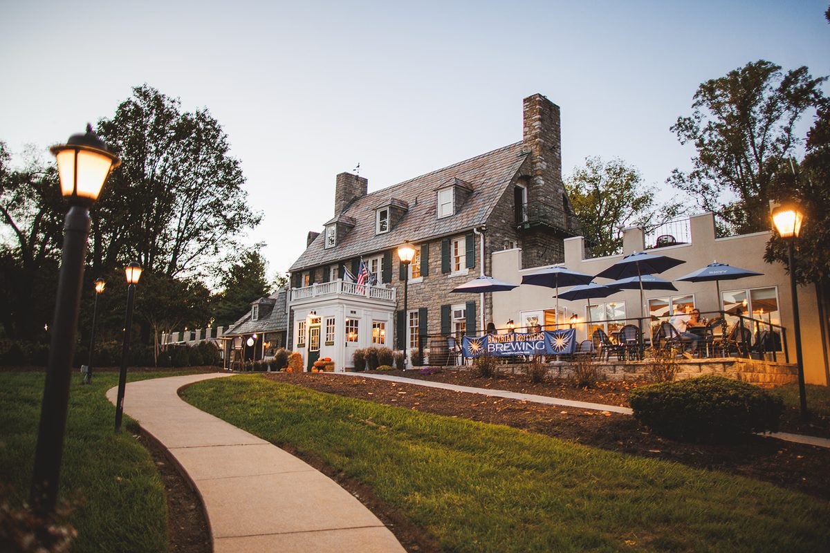 The stone and slate main house of the Bavarian Inn at dusk, lamps glowing along the curving path and guests seated under umbrellas on the terrace