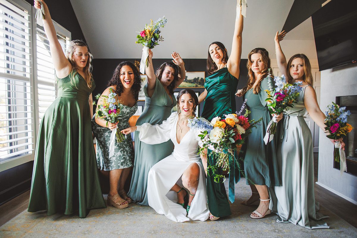 Andrea kneeling at the center of her bridal party as all of them raise bouquets and arms in a cheer, every shade of green around her