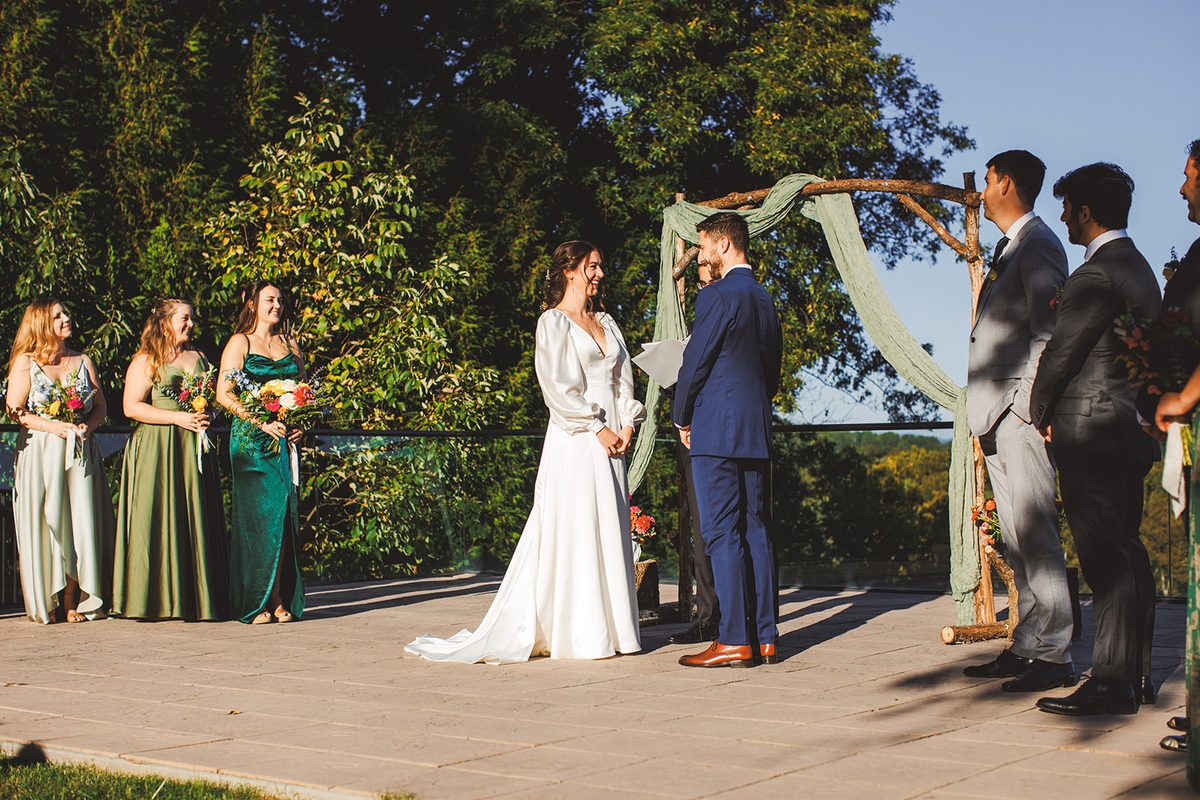 Andrea and Patrick facing each other at the sage-draped arch on the terrace, bridesmaids in greens to one side and groomsmen to the other