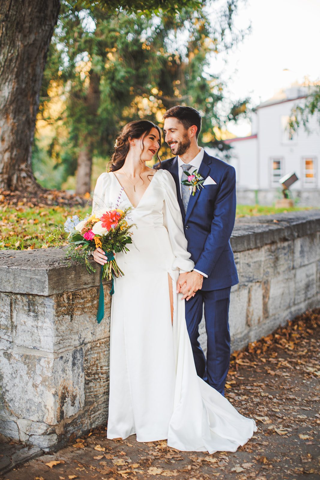 Andrea and Patrick standing hand in hand along a low stone wall in golden light, turned toward each other, her bright bouquet at her side