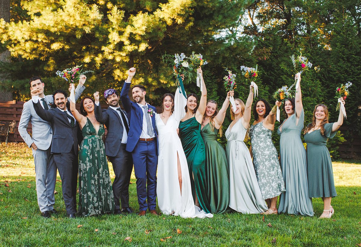 The full wedding party in navy, gray, and a dozen shades of green cheering with bouquets raised on the lawn