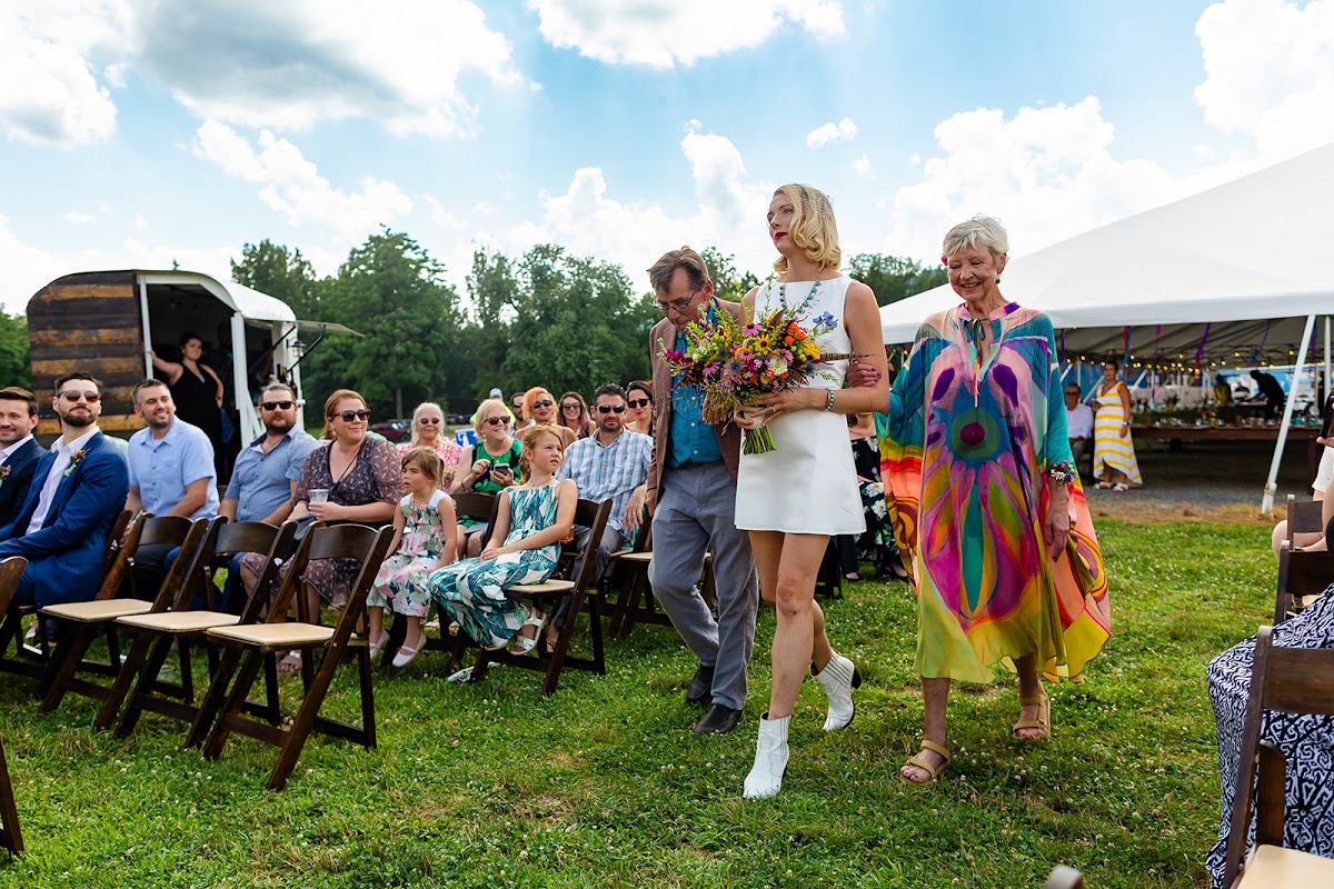 Annie walking to the ceremony with her wildflower bouquet, escorted across the lawn past standing guests, the reception tent behind