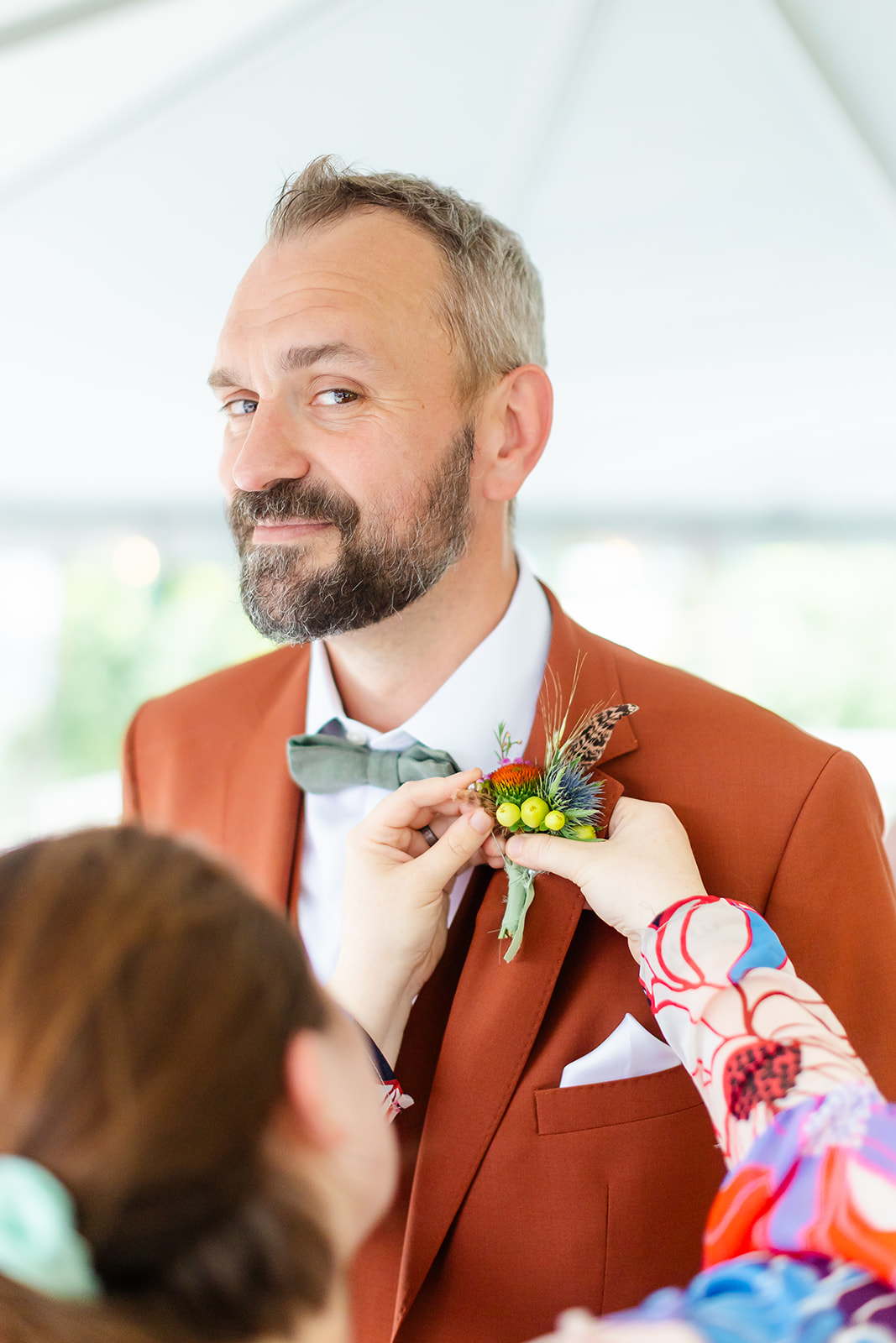 Thomas grinning as a boutonniere of coneflower, thistle, berries, and a feather is pinned to the lapel of his rust suit under the tent