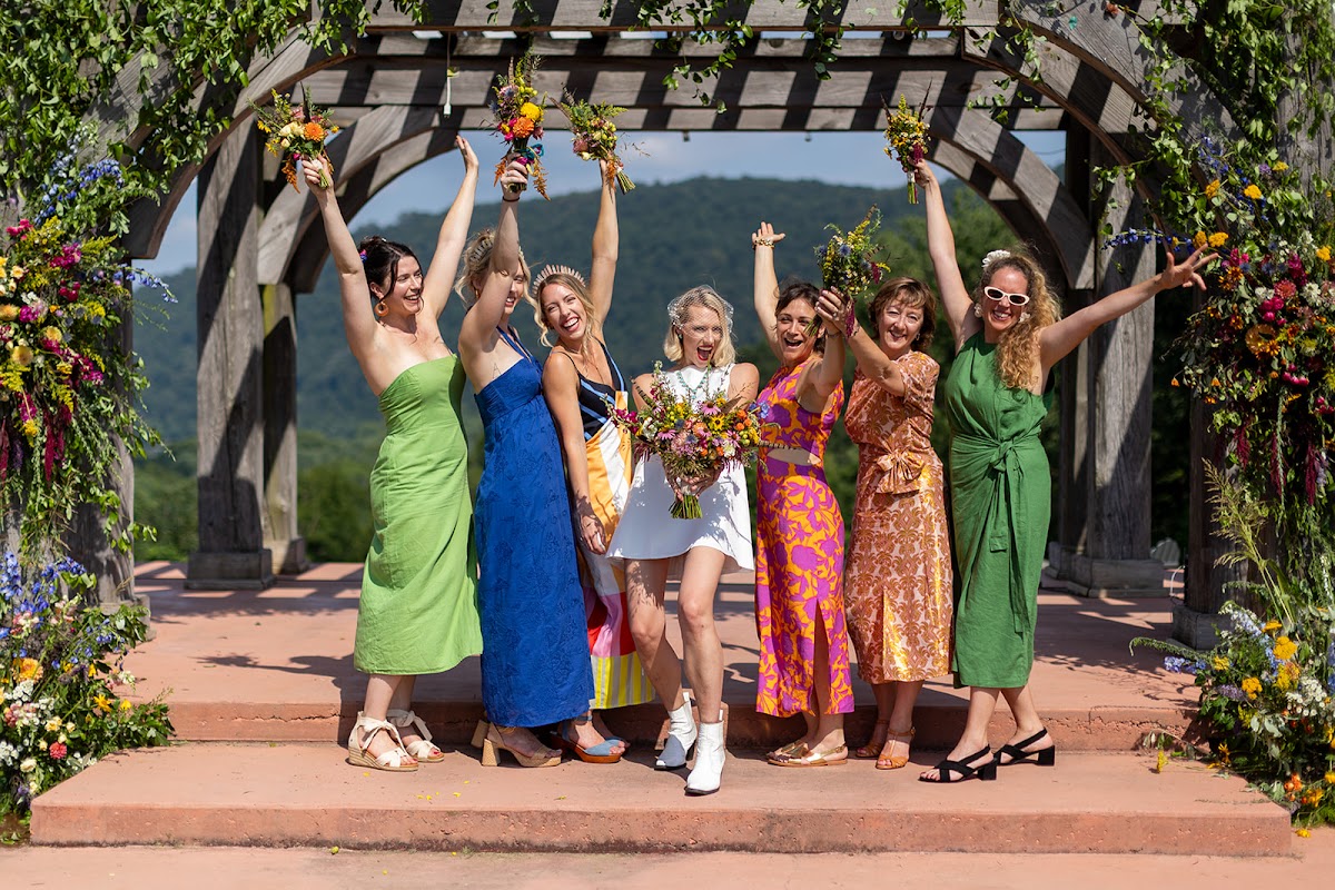 A wedding party cheering with bouquets raised under a flower-draped pergola, mountains behind them