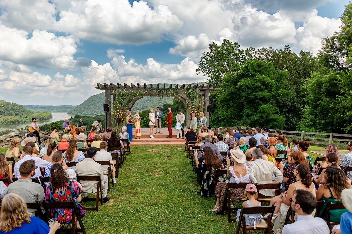 Wide view of the ceremony at Harpers Ferry Brewing: guests flank a grass aisle leading to the timber pergola, with the Potomac and wooded ridgelines under a blue summer sky