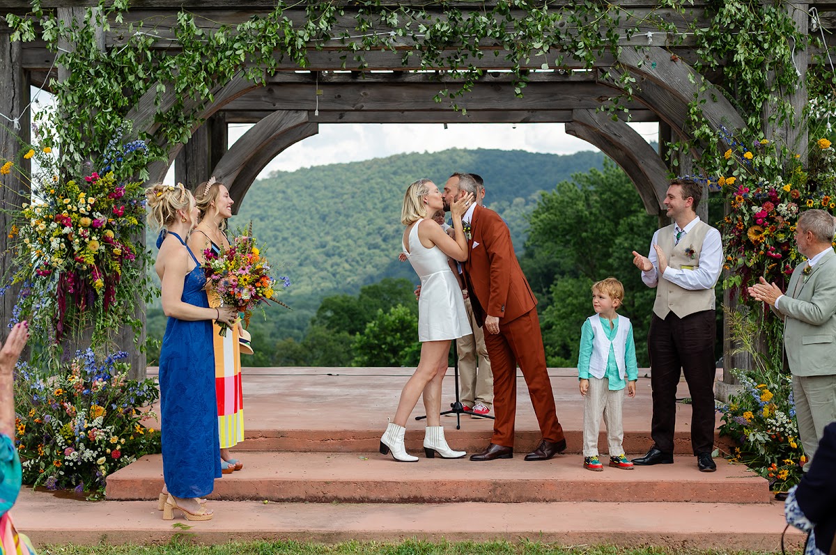 A first kiss under a vine-wrapped pergola above the river as the wedding party reacts on both sides