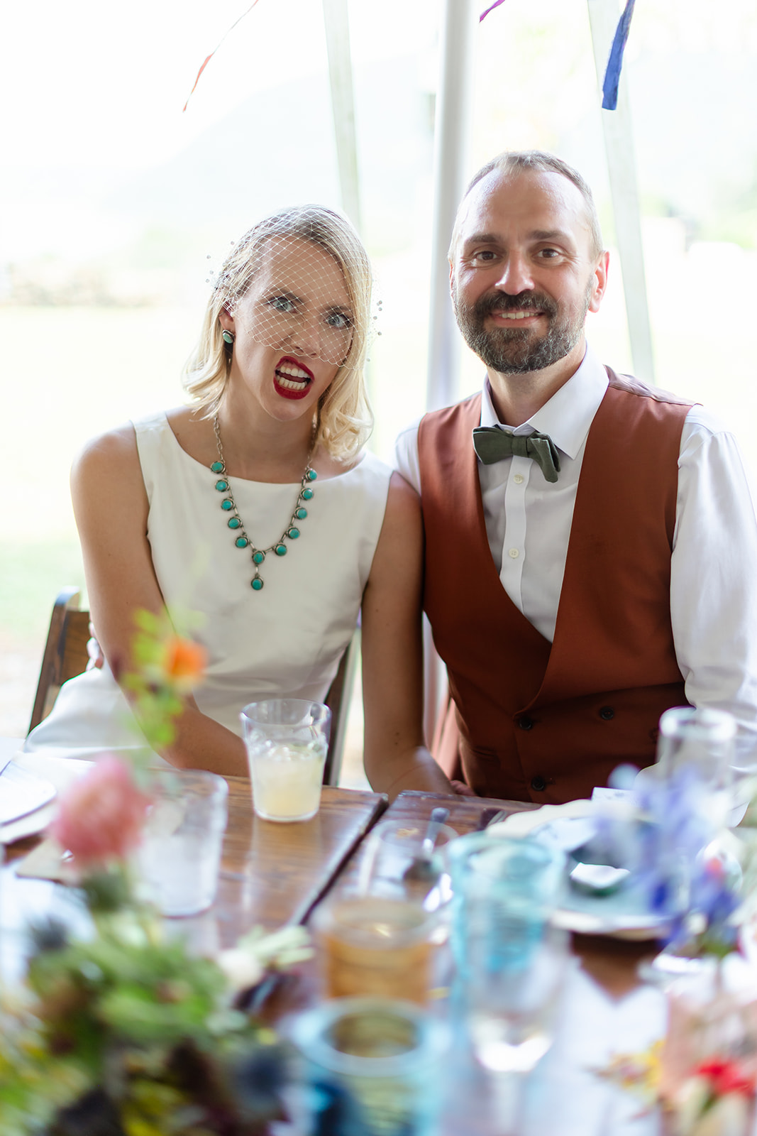 Annie snarling playfully at the camera in her birdcage veil and turquoise necklace while Thomas smiles beside her at the head table