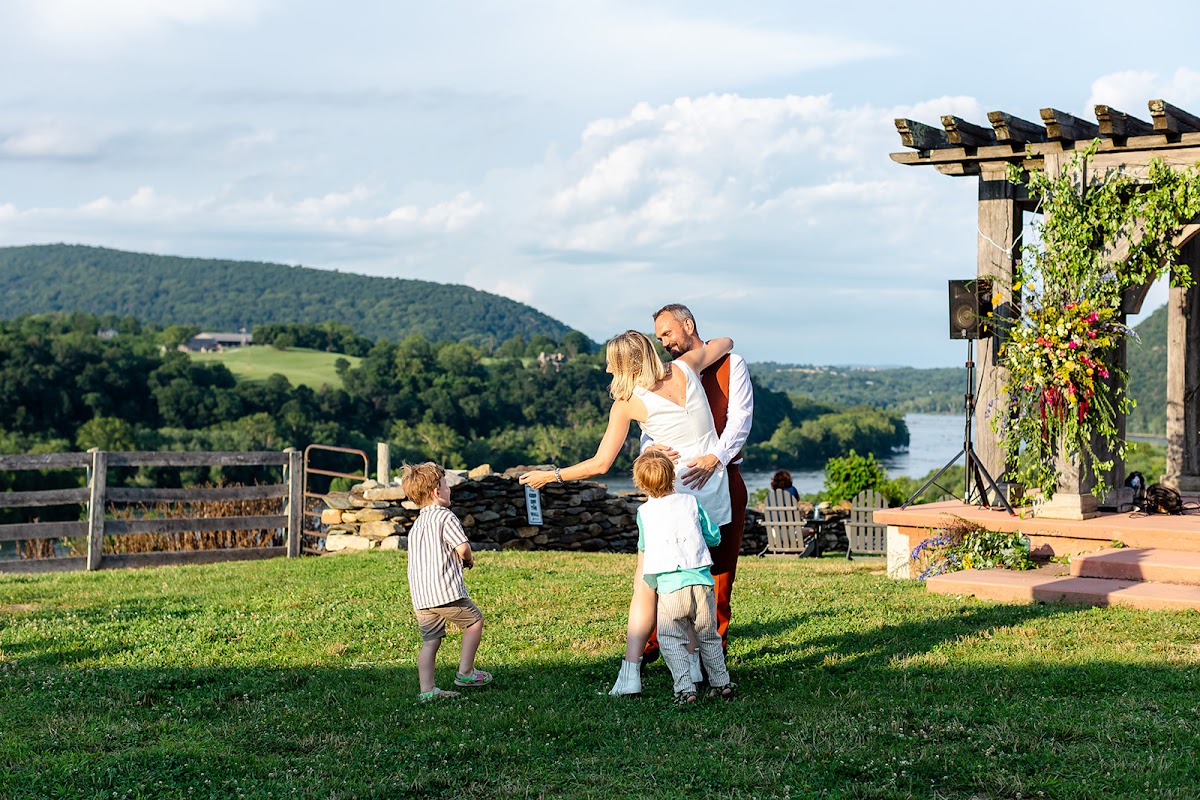 Annie and Thomas embracing on the lawn near the pergola while two small kids orbit them, the river valley glowing in evening light