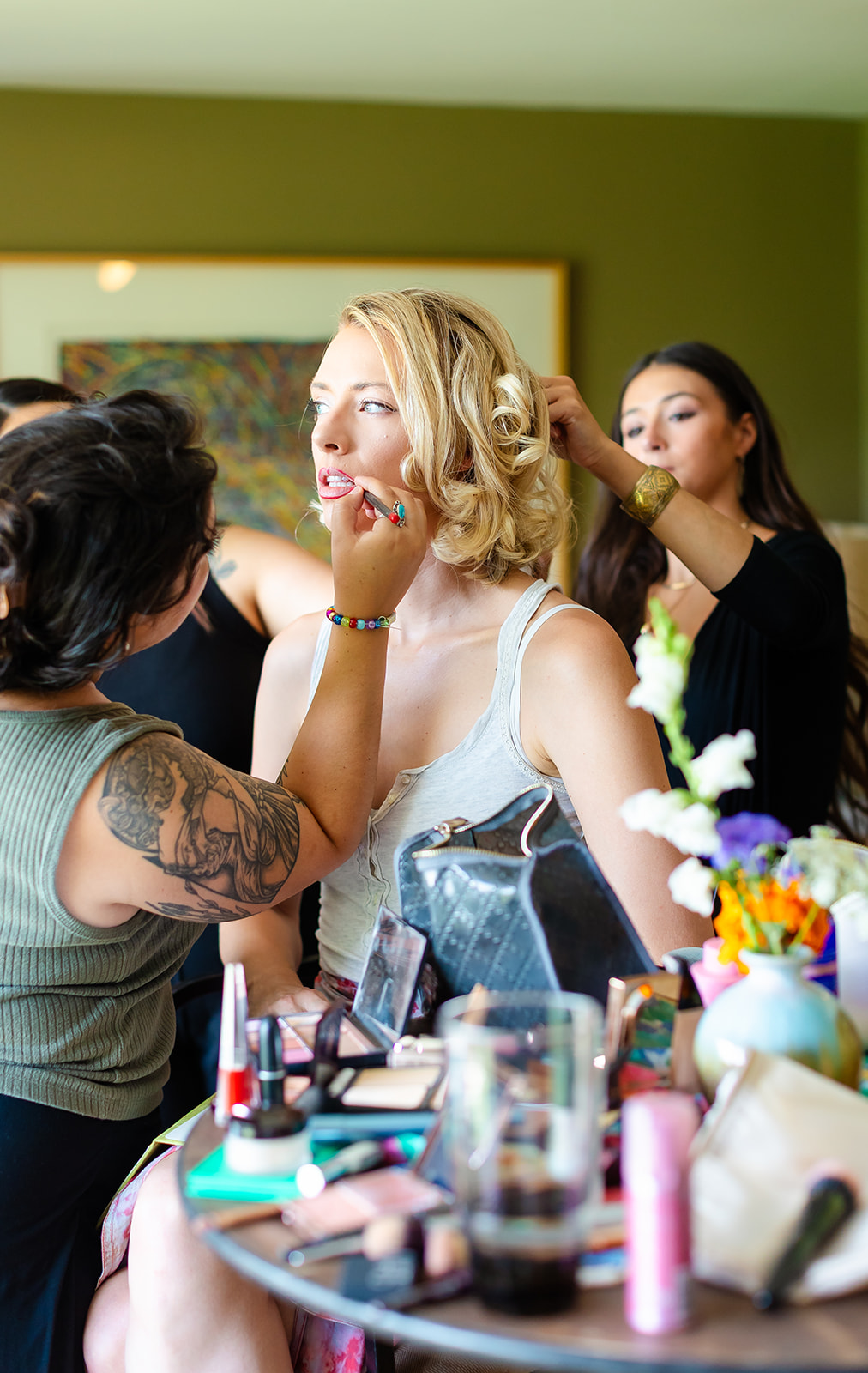 Annie in the makeup chair having her lip color finished while two artists work, snapdragons and flowers on the table in front of her