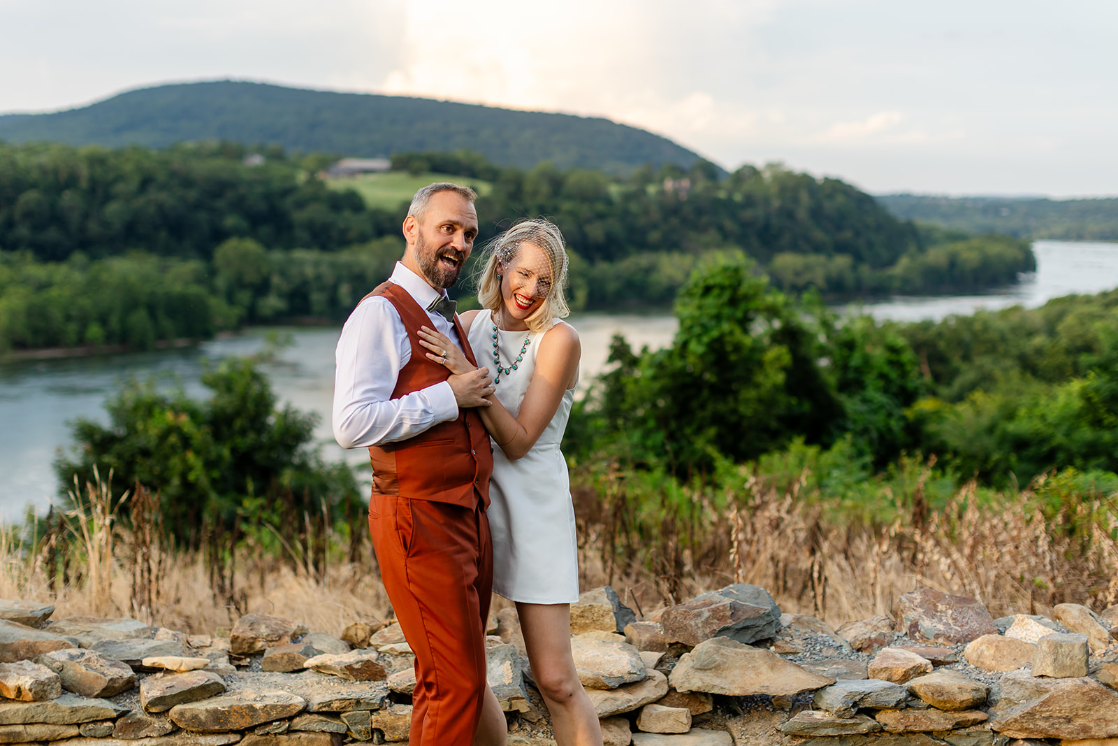 Annie laughing into Thomas at the stone overlook wall at Harpers Ferry Brewing, the Potomac curving through wooded hills behind them