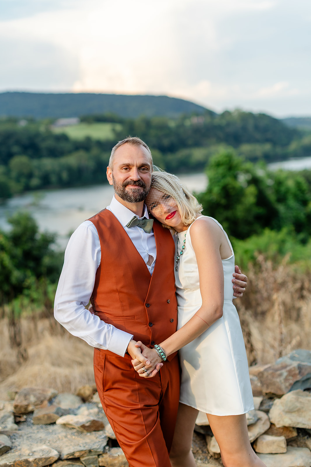 Annie resting her head on Thomas's shoulder at the overlook, hands clasped, the river valley in soft evening light behind them