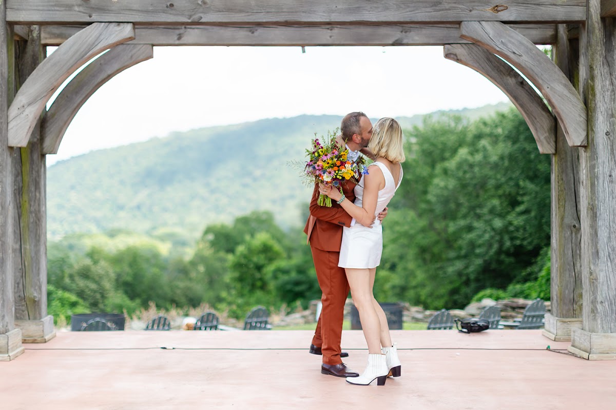 Annie and Thomas embracing on the stage under the timber pergola arch, framed against a wooded green mountain