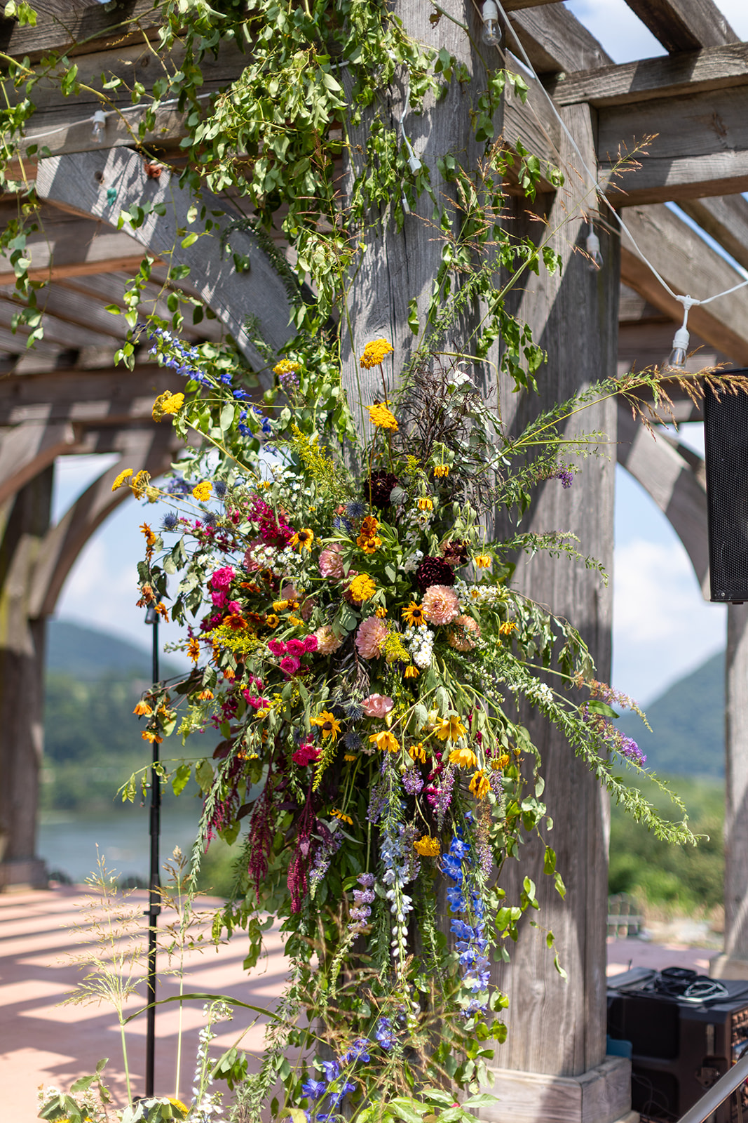 A wildflower installation in pinks, yellows, and blues climbing a timber pergola post, with string lights and the hazy ridge behind