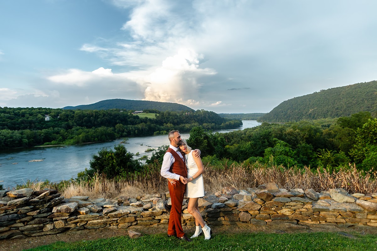A couple holding each other on a river overlook beneath a towering sunlit cloud