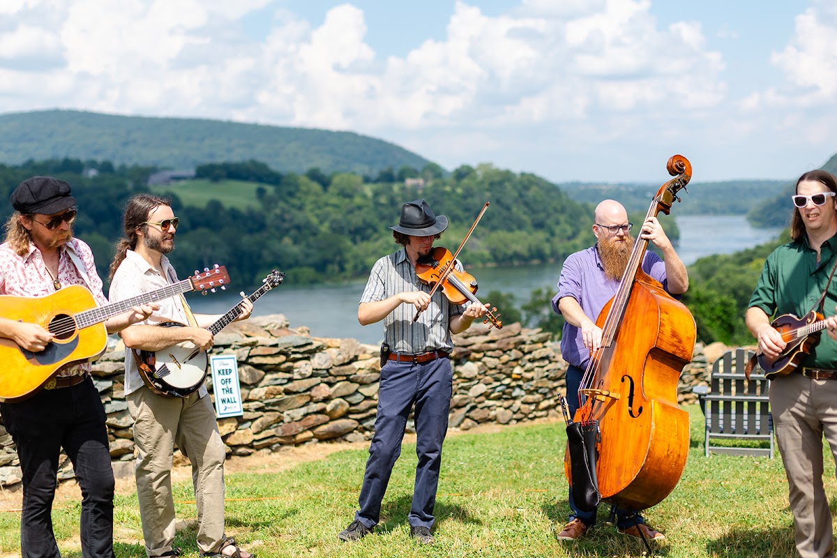 A five-piece string band with guitars, banjo, fiddle, and upright bass playing on the lawn beside the overlook wall, the Potomac behind them