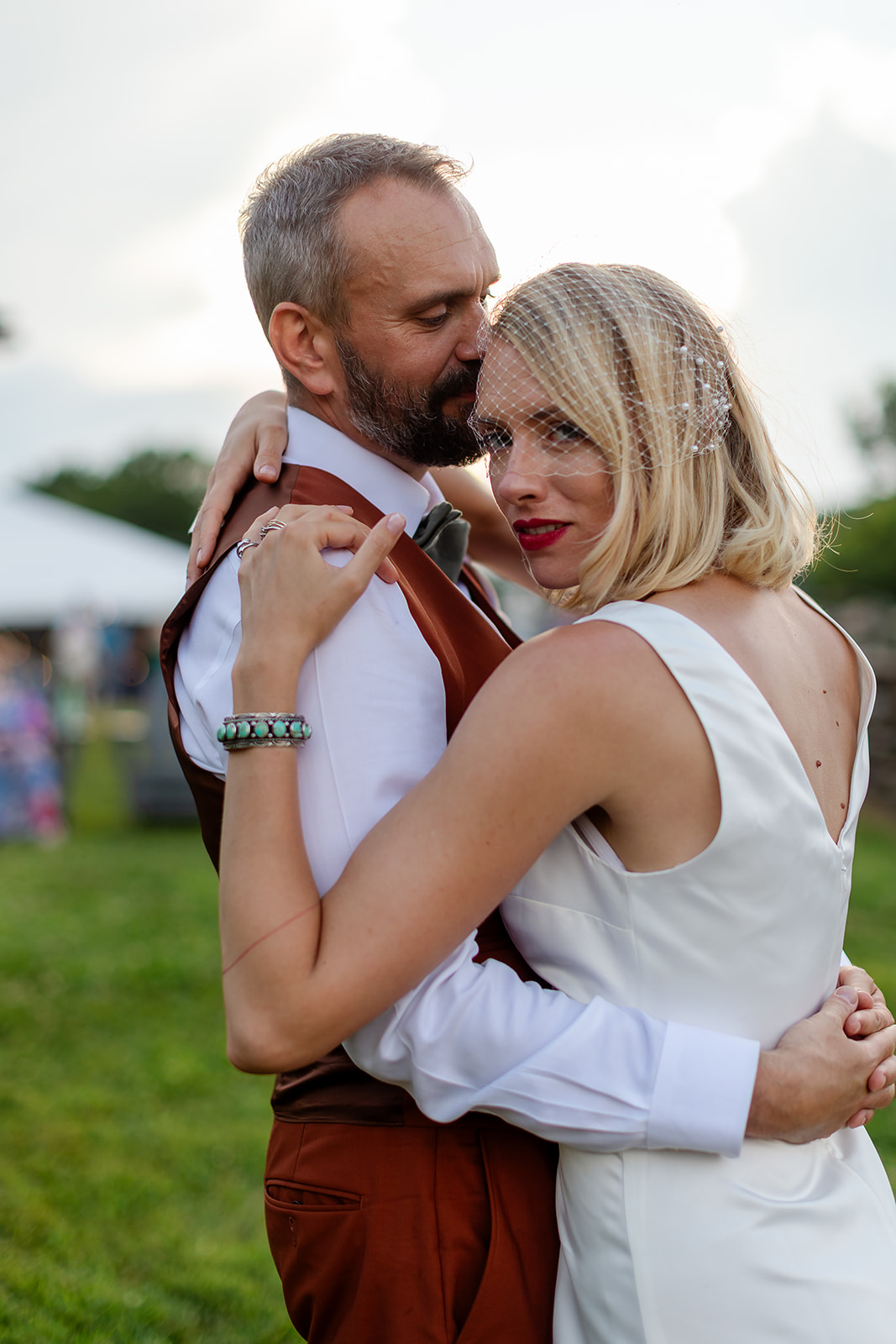 Thomas pressing a kiss to Annie's temple on the lawn, her birdcage veil and red lip catching the light, the reception tent soft in the background