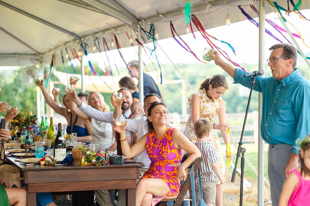 Guests under the ribbon-strung tent raising glasses during a toast, a man at the microphone lifting a mason jar