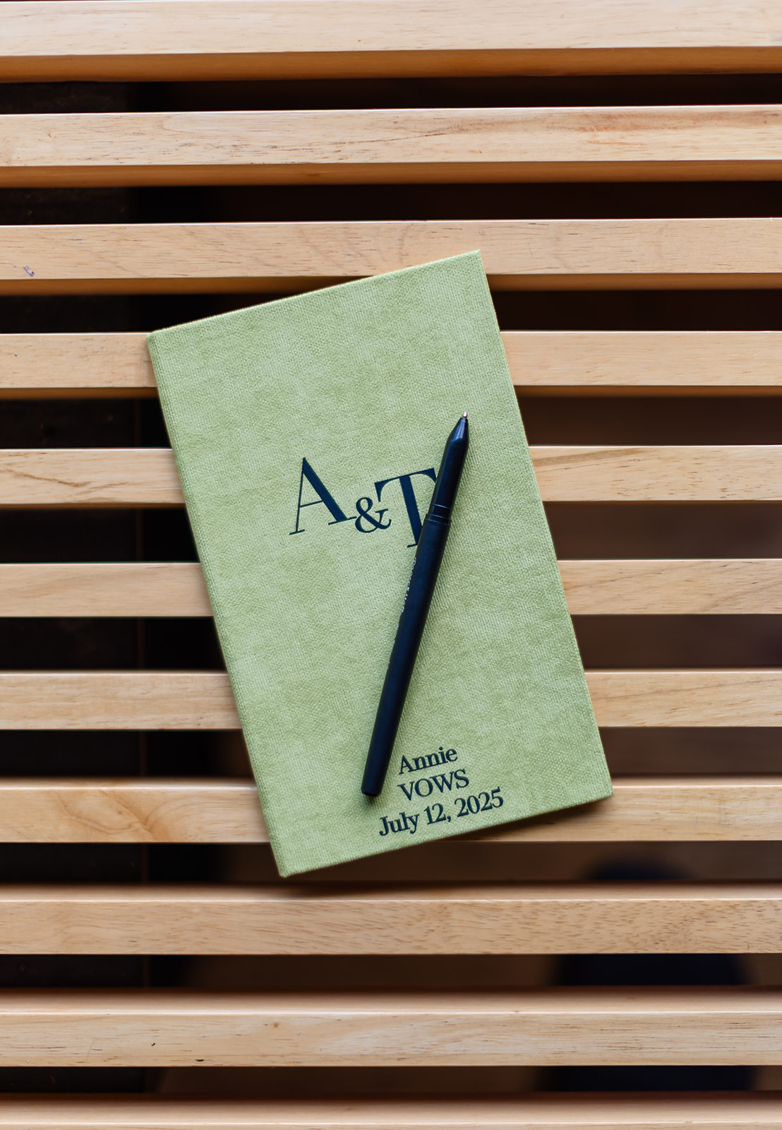 A green vow book stamped A and T with a pen resting on it, printed Annie, Vows, July 12, 2025, lying on a slatted wooden bench
