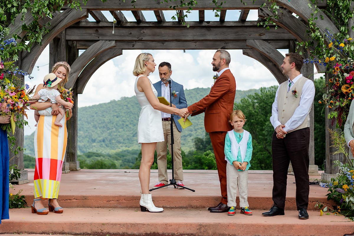 Annie and Thomas holding hands during vows on the pergola stage at Harpers Ferry Brewing, the officiant at the microphone and the wedding party beside them, green mountains behind