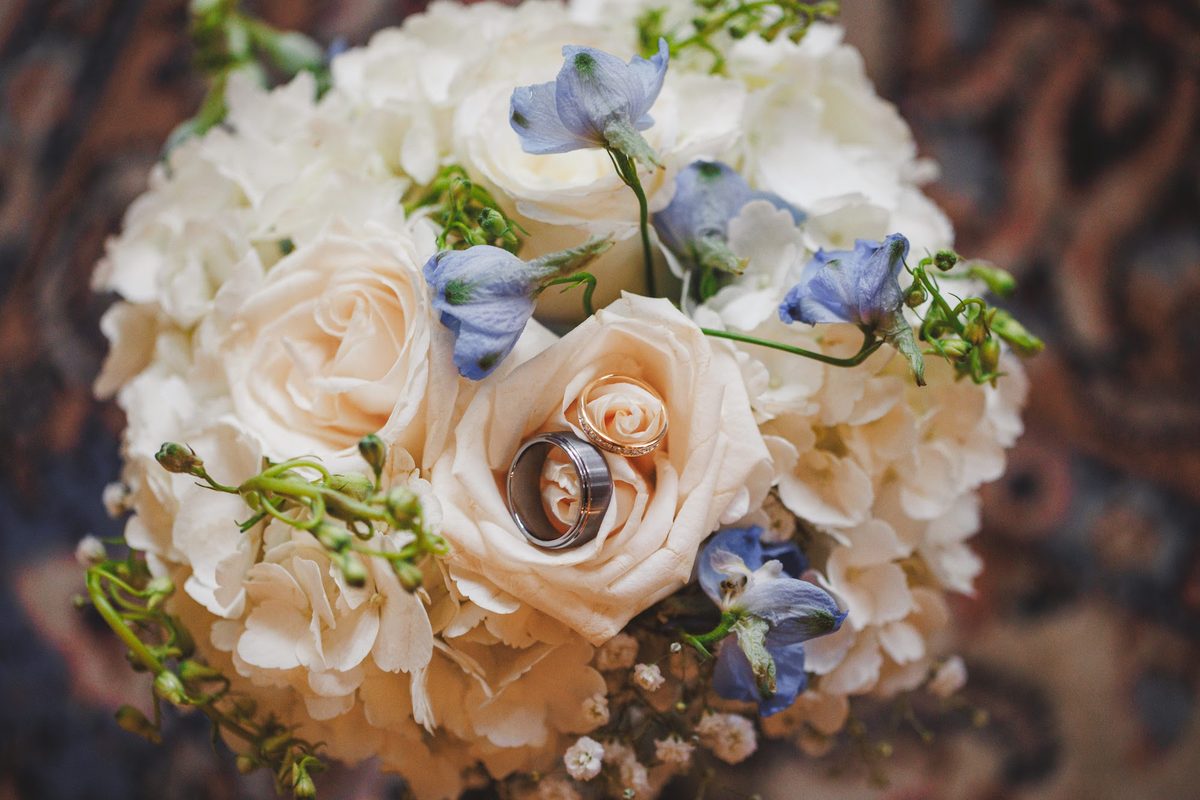Both wedding bands nestled into a cream rose at the center of a bouquet of white hydrangea and pale blue delphinium
