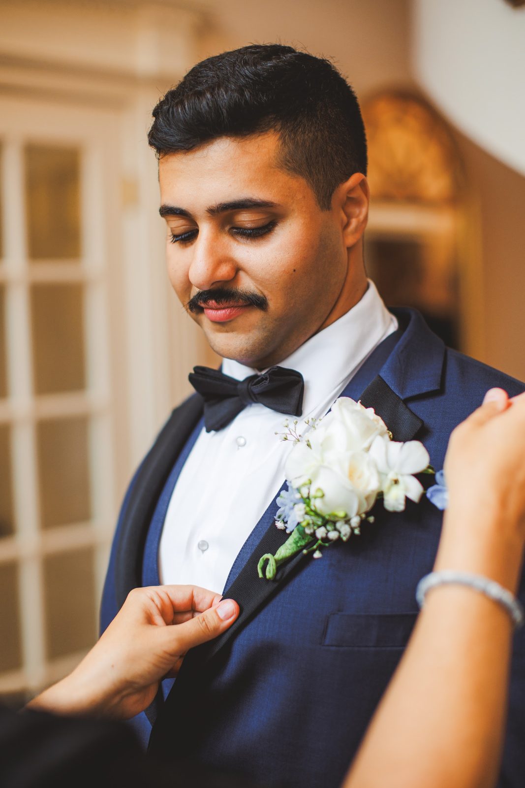 Youssef looking down with a small smile as a white rose boutonniere with pale blue accents is pinned to his navy tuxedo lapel