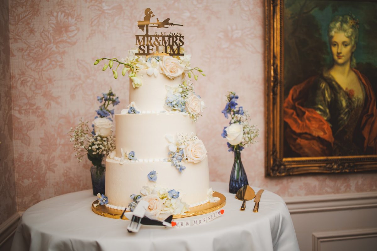 Three-tier white wedding cake with blue delphinium and cream roses, a gold topper reading Mr and Mrs Selim with the date 8.31.2024 and a small airplane