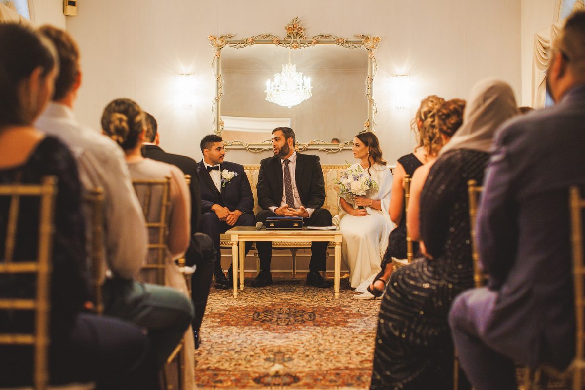 The Islamic marriage ceremony in the estate's sitting room, Bailey and Youssef seated with the officiant beneath a crystal chandelier and gilt mirror, guests close on gold chairs