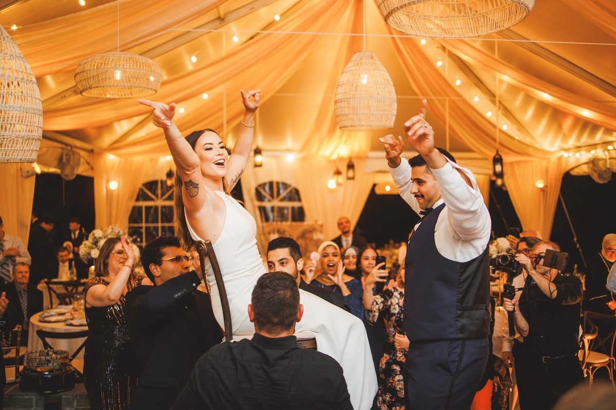 Bailey lifted on a chair with both arms in the air while Youssef dances beside her, drums and cheering guests around the glowing tent